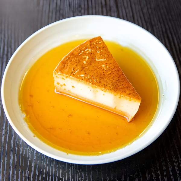 A slice of Cuban flan with caramel sauce in a white bowl on a dark wooden table.
